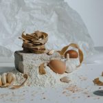 A creative still life featuring pasta, eggs, flour, and garlic with a rustic kitchen vibe.