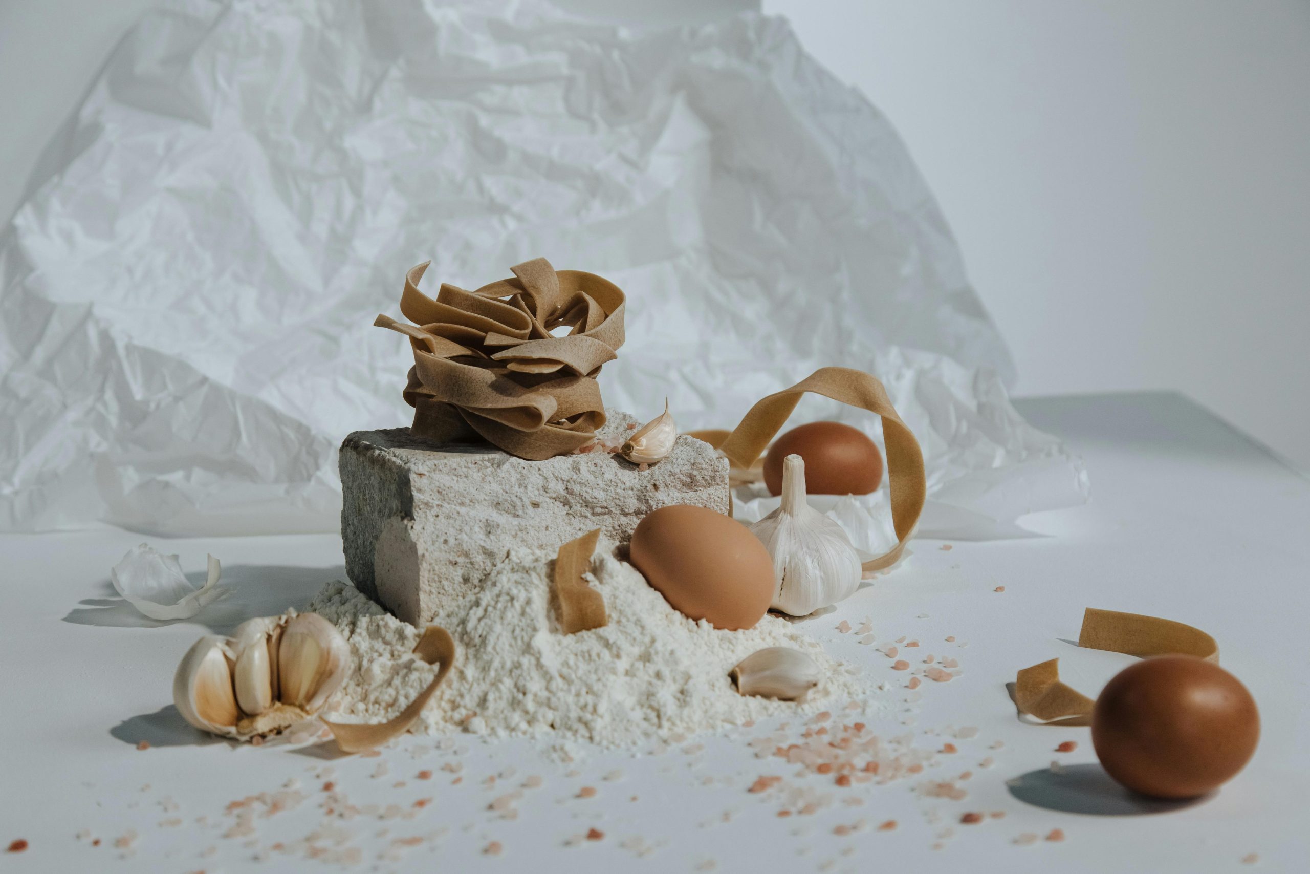 A creative still life featuring pasta, eggs, flour, and garlic with a rustic kitchen vibe.