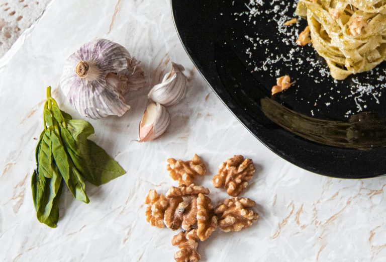 Flat lay of pesto pasta, basil, garlic, and walnuts on a marble surface for Italian cuisine.